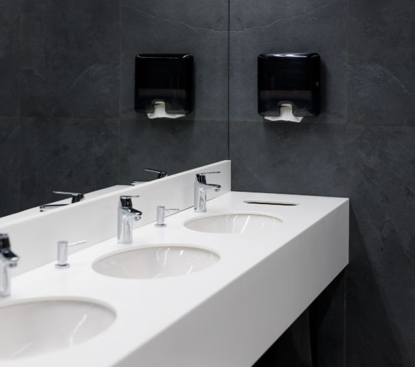 Modern public restroom sinks with soap dispensers on a dark wall.