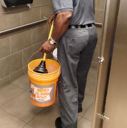 Man holding a plunger and a Home Depot bucket in a restroom.